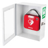 A white wall‑mounted cabinet with an open clear door holding a red Philips HeartStart defibrillator case featuring a heart and lightning bolt icon. A green AED sign is visible on the cabinet.