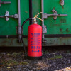 Red p50 6kg powder fire extinguisher standing on gravel in front of a green work site container