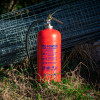 Red p50 6kg powder fire extinguisher standing on grass near a metal work site mesh fence