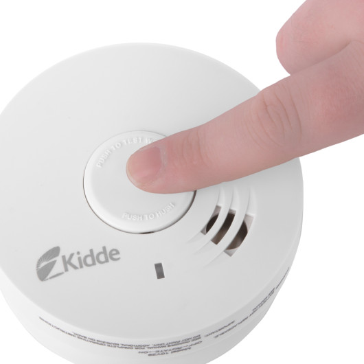 Finger pressing the test button of a white Kidde Ke10Y29 Compact Design smoke alarm