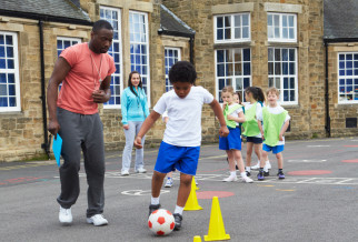 School children playing football outdoors with a male coach supervising, related to Defibrillators For Schools