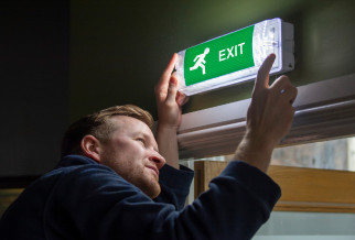 Man testing illuminated emergency lighting exit sign on wall titled Emergency Lighting Testing