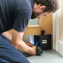 Engineer installing dorgard smartsound door retainer with a drill at the base of an interior wooden door