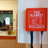 Ultrafire fire blanket in red plastic case hanging on a white kitchen cupboard with two black straps visible