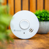 White Ke10Y29 Kidde 10Y29 10 Year Optical Smoke Alarm placed on a wooden surface with blurred plants in the background