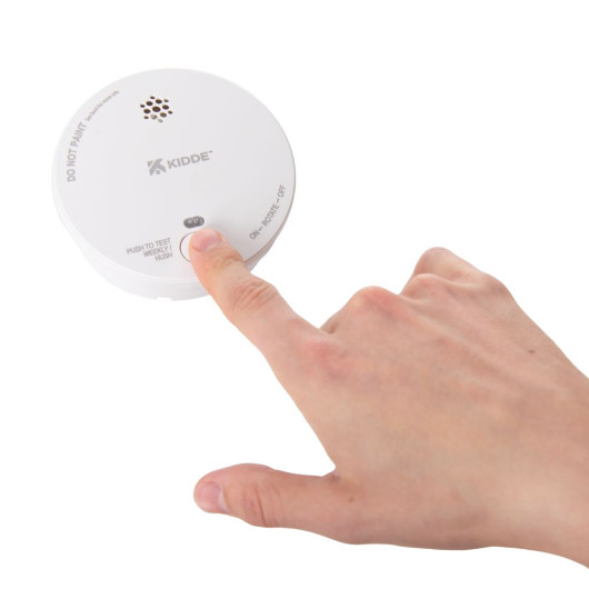 A close-up of a hand pressing the test button on a white Kidde smoke alarm, showing the front vent, brand logo, and “Push to Test Weekly / Hush” label.