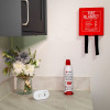Wall‑mounted red fire blanket with pull straps above a Kidde Flame Out fire‑fighting spray and a white UltraFire alarm on a kitchen countertop beside a vase of artificial flowers