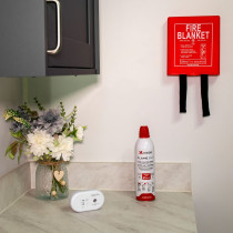 Wall‑mounted red fire blanket with pull straps above a Kidde Flame Out fire‑fighting spray and a white UltraFire alarm on a kitchen countertop beside a vase of artificial flowers