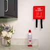 Wall‑mounted red fire blanket with pull straps above a Kidde Flame Out fire‑fighting spray on a kitchen countertop beside a vase of artificial flowers