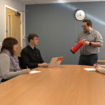 People attending fire safety awareness training in office with a man holding a red fire extinguisher near a table