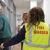 A fire warden wearing a high‑visibility vest guides people through a corridor during an evacuation drill.
