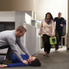 A woman carrying a Zoll Aed 3 Defibrilator running through an office towards a person lying on the floor receiving CPR from a man