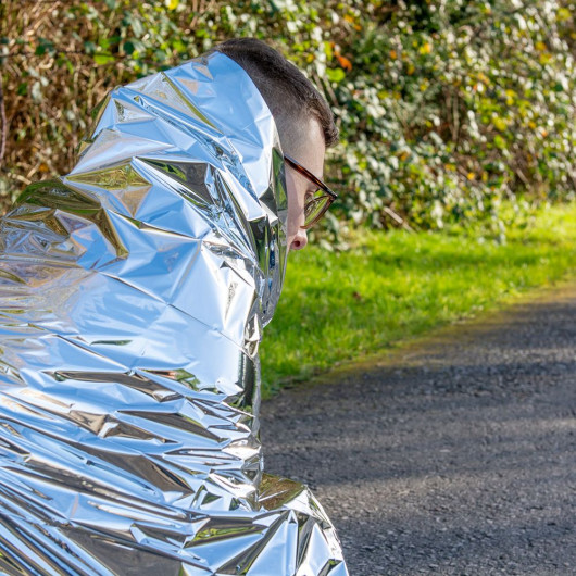 A person sits on the edge of a paved path, wrapped in a reflective silver emergency blanket. Green bushes and foliage fill the background, and the crinkled thermal material covers most of their body, suggesting they are keeping warm outdoors.