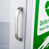 Close‑up of a white cabinet door with a metal handle and partial green “Defibrillator / Heart Res…” label.